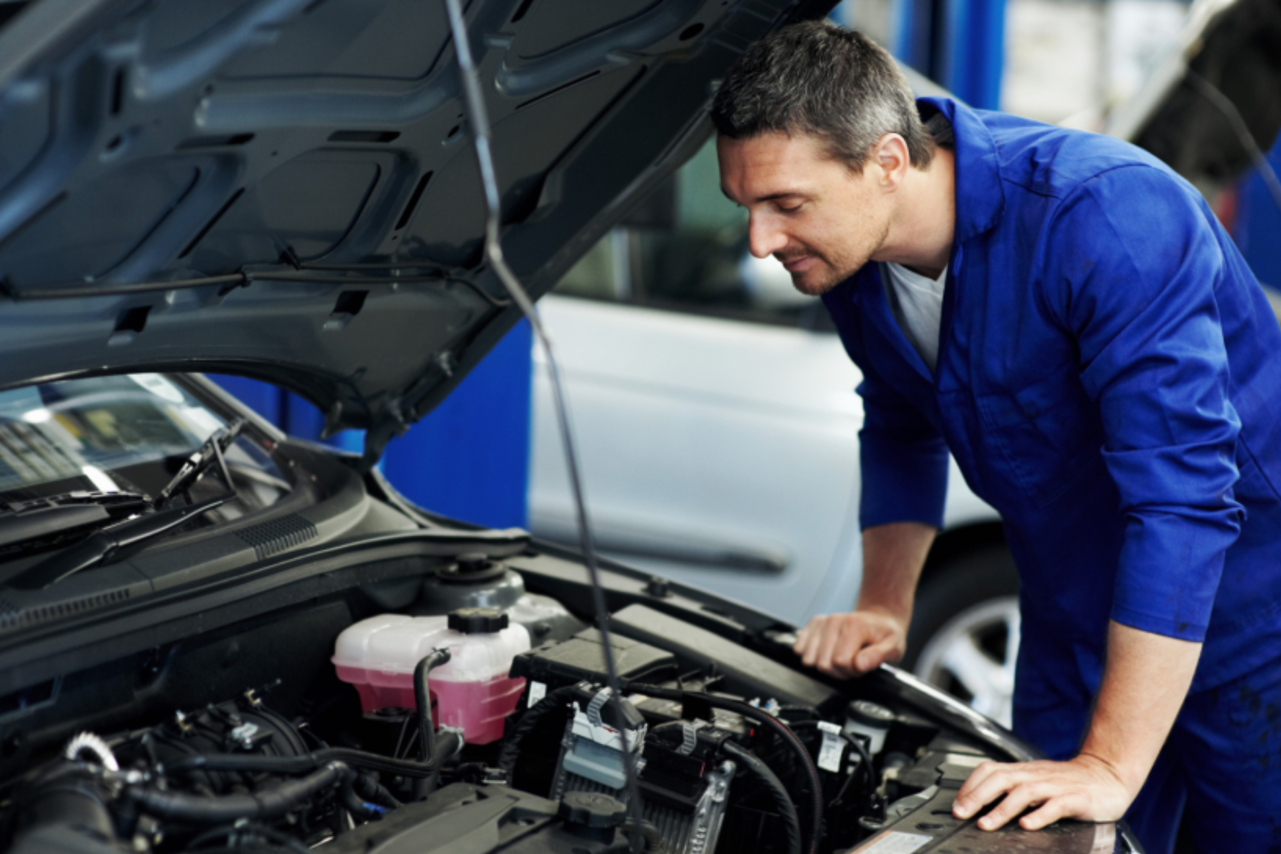 Mechanic in blue coveralls leaning over an open car hood, inspecting the vehicle's engine bay and fluid reservoirs.